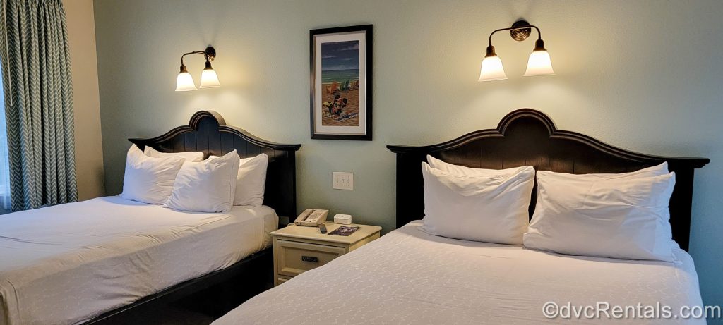 The two queen beds in the Deluxe Studio at Disney’s Old Key West Resort. Between the beds is a framed picture of Minnie Mouse at the beach, and the room is decorated with light neutrals and pastels.