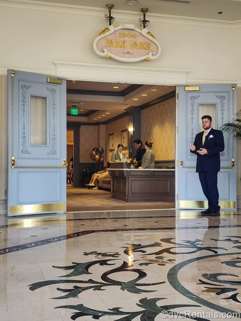 Cast Members await guests at the entry way into 1900 Park Fare at Disney’s Grand Floridian Resort & Spa. Above the blue doors is a pink and gold sign displaying the restaurant’s name.