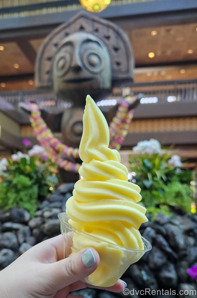 A hand holds up a cut of pineapple Dole Whip in the lobby of Disney’s Polynesian Villas & Bungalows. A tiki statue and landscaping can be seen in the background.