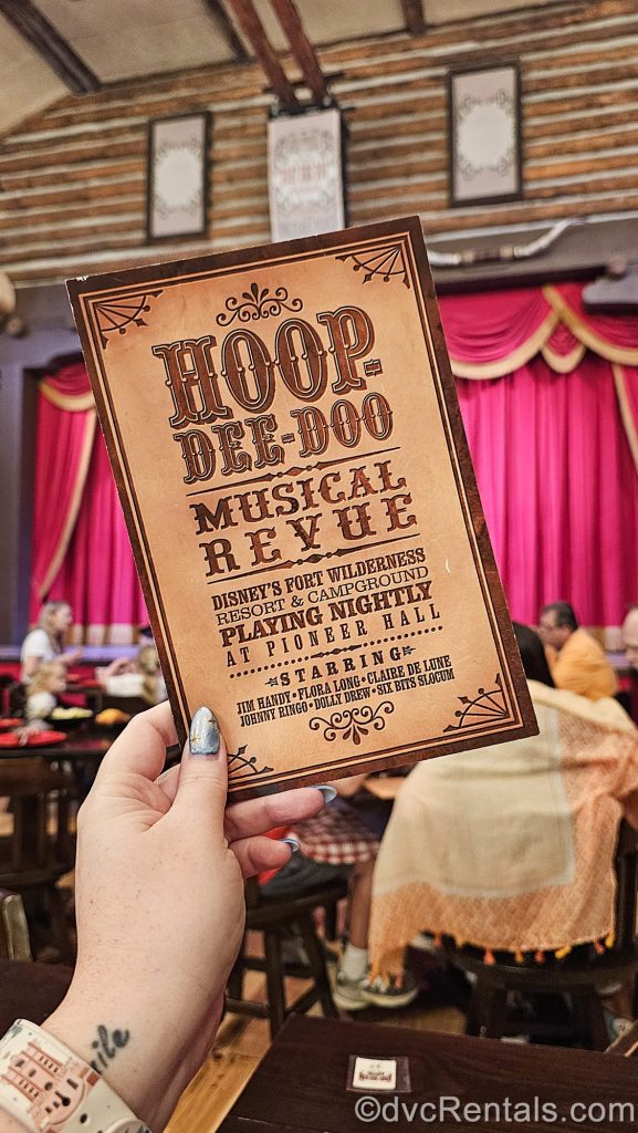 A hand holds a brown card reading “Hoop-Dee-Doo Musical Revue” along with details about the dinner show while the restaurant’s interior can be seen in the background.