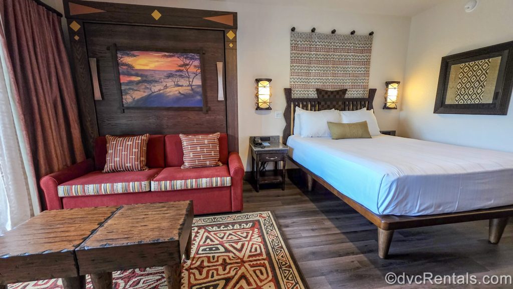 A Deluxe Studio at Disney’s Animal Kingdom Villas Kidani Village. The room is decorated with dark woods, neutral paint, and bright pops of orange and red in the African-inspired art and Lion King touches.